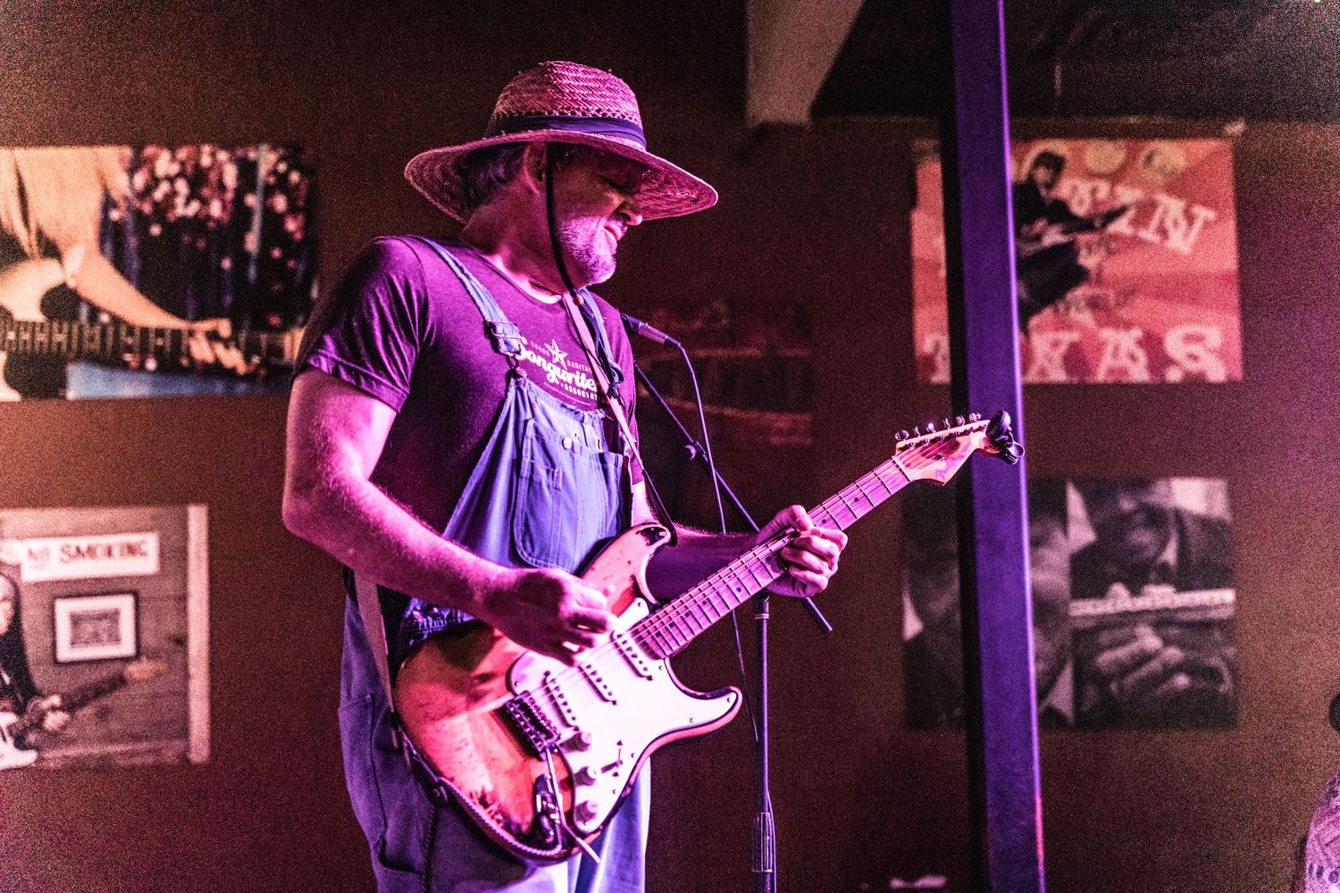 Guitarist wearing a purple hat performs on stage with vintage photos displayed on the wall behind.
