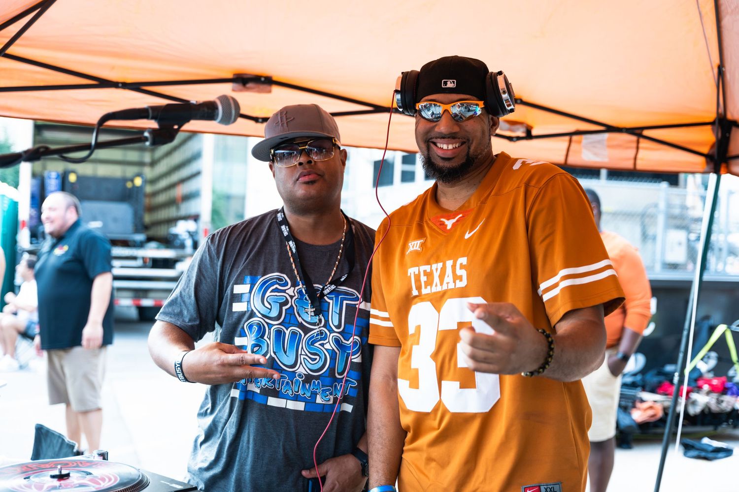 Two people posing together at outdoor event wearing sports jerseys and sunglasses.