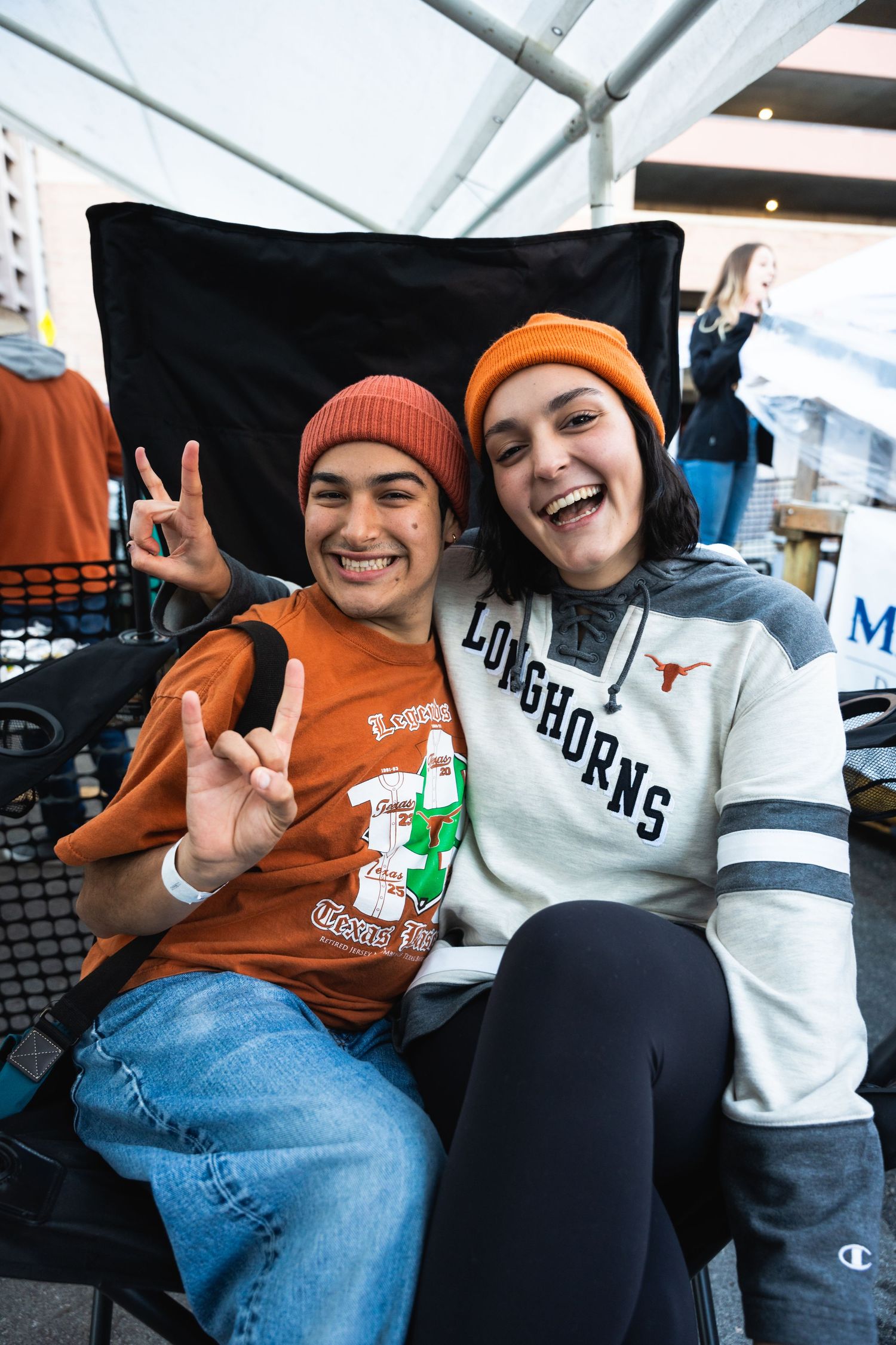 Two friends in orange and white beanies making peace signs while enjoying a sporting event together.