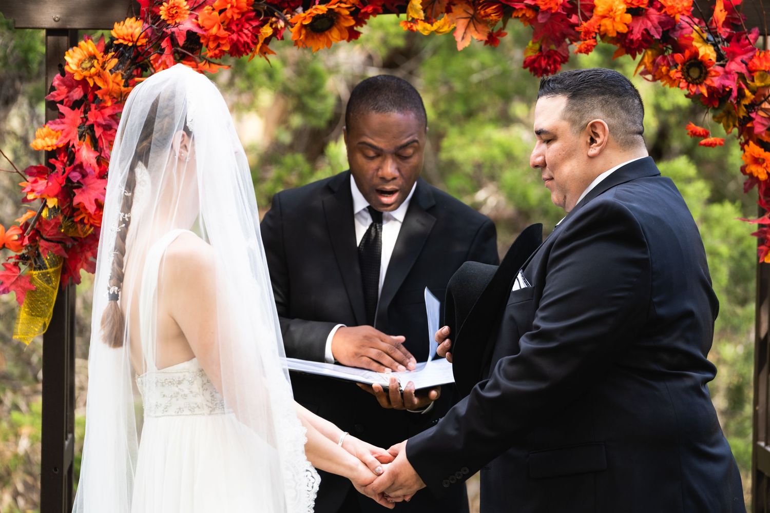 A wedding ceremony moment under a bright autumn-colored floral arch during an outdoor service.
