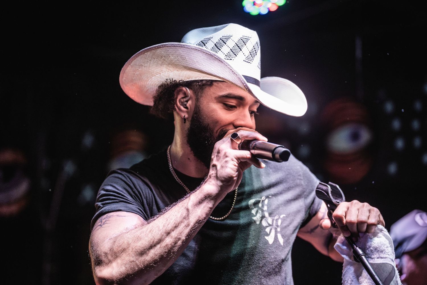 Country singer performs on stage wearing a white cowboy hat and black t-shirt while gripping microphone.