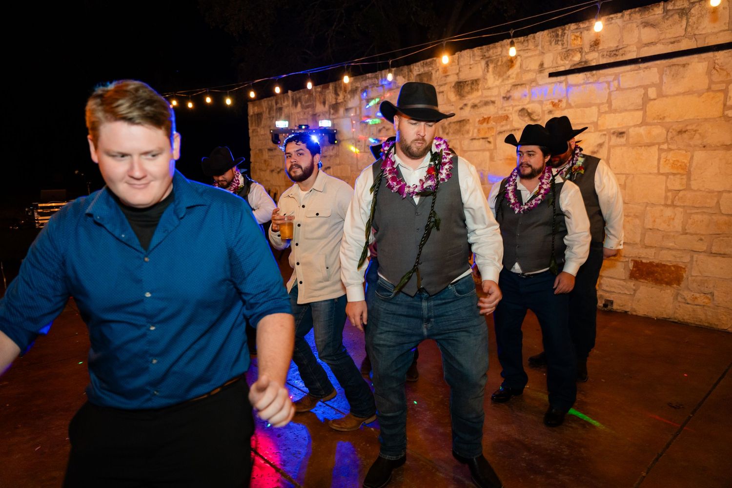 A group of men in cowboy hats and vests dance in formation under string lights at night.