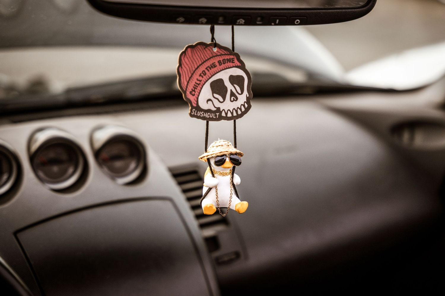 Decorative skull ornament and small figure hanging from rearview mirror in car interior.