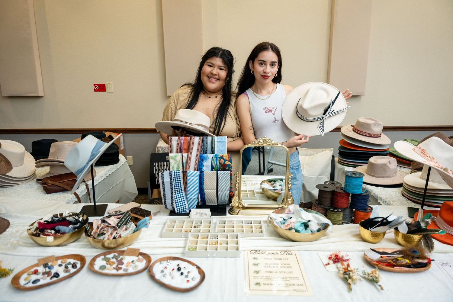 Two vendors standing behind table display of food and merchandise at an event.