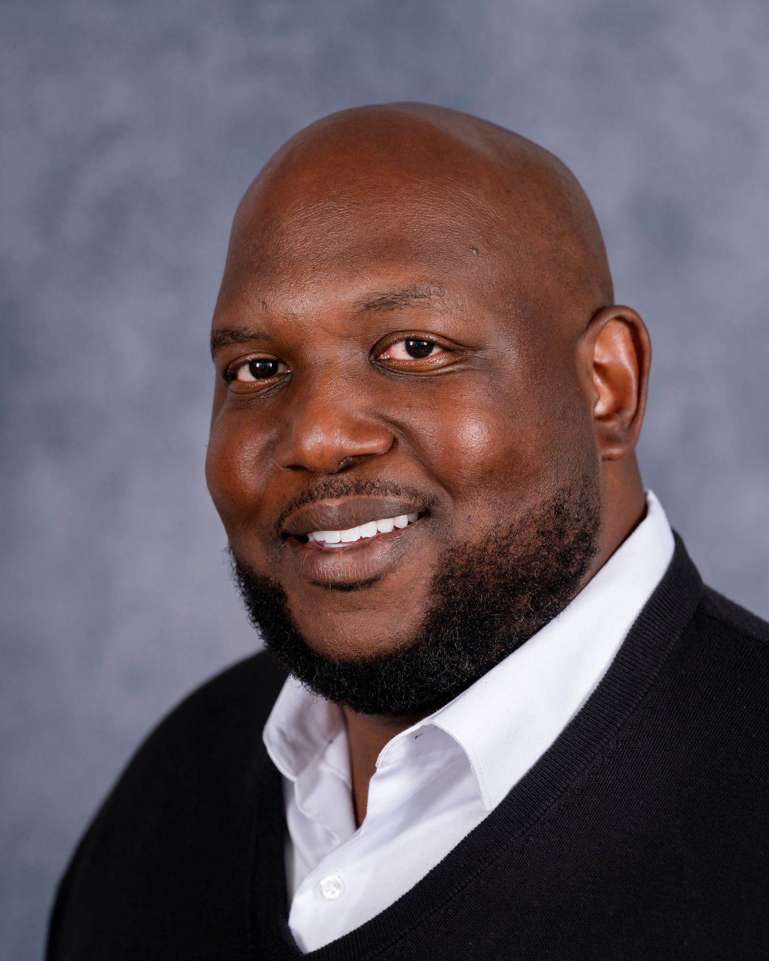 Professional headshot of a person wearing a black blazer and white dress shirt smiling warmly against a gray backdrop.