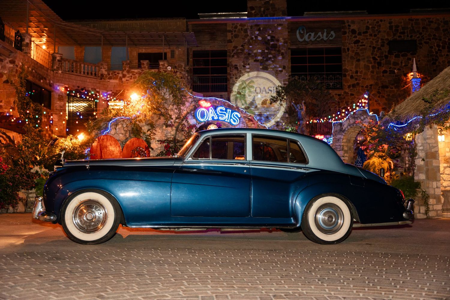 A classic blue vintage car with white wall tires parked against a festively lit building.
