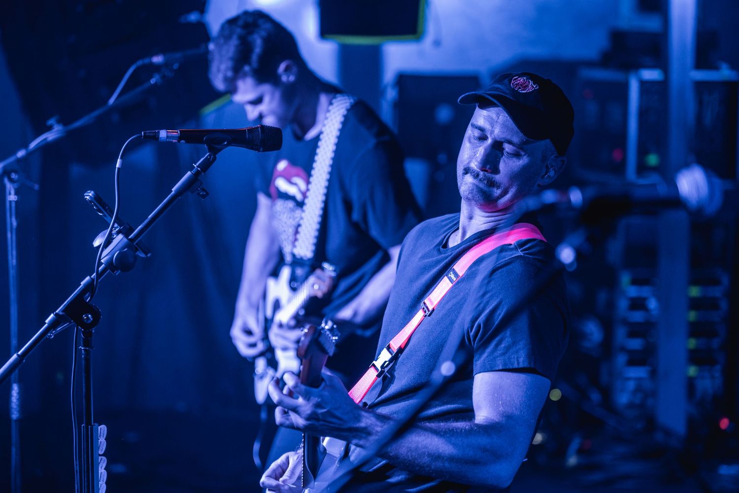 Musicians perform on stage with blue lighting and guitar equipment visible.