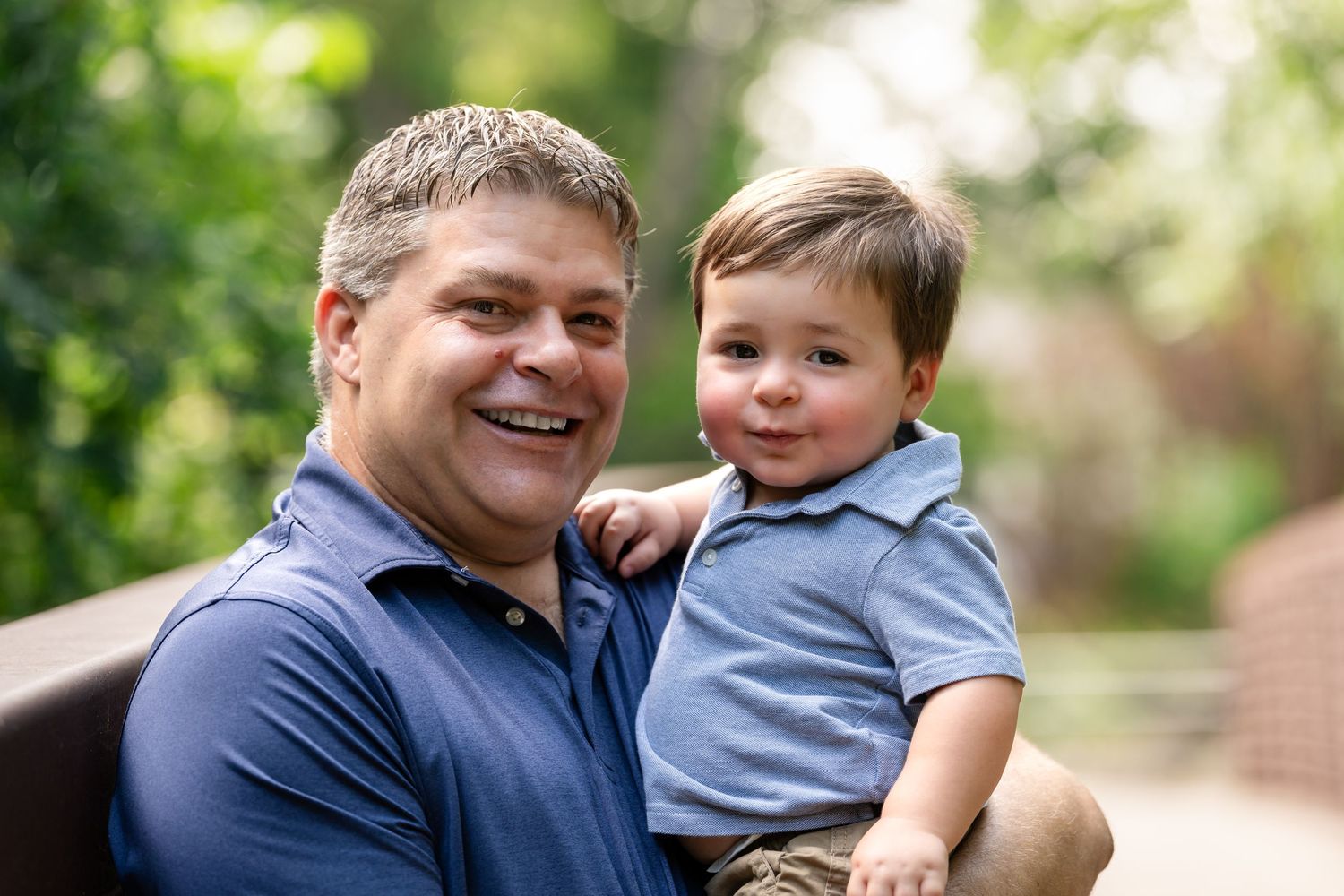 A toddler in a blue polo shirt sitting close with an adult on a summer day.