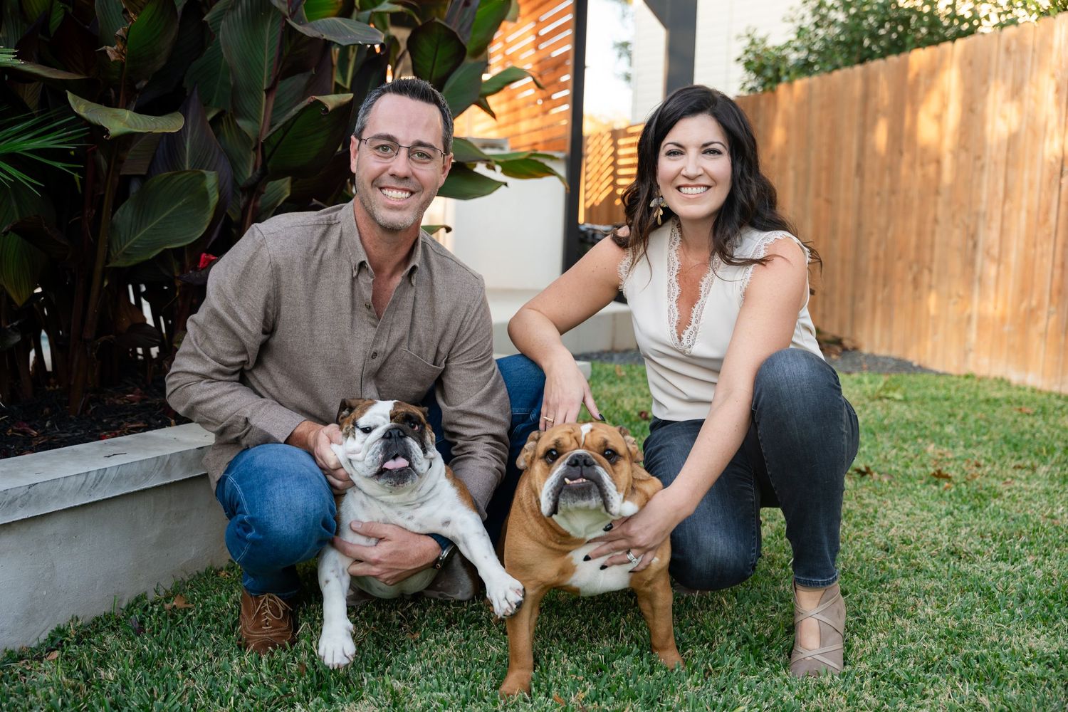 A couple poses with their two dogs in a backyard garden with tropical plants and a wooden fence.