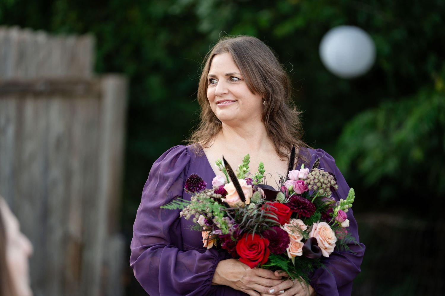 Wedding attendant in purple dress holds colorful floral bouquet during outdoor ceremony.