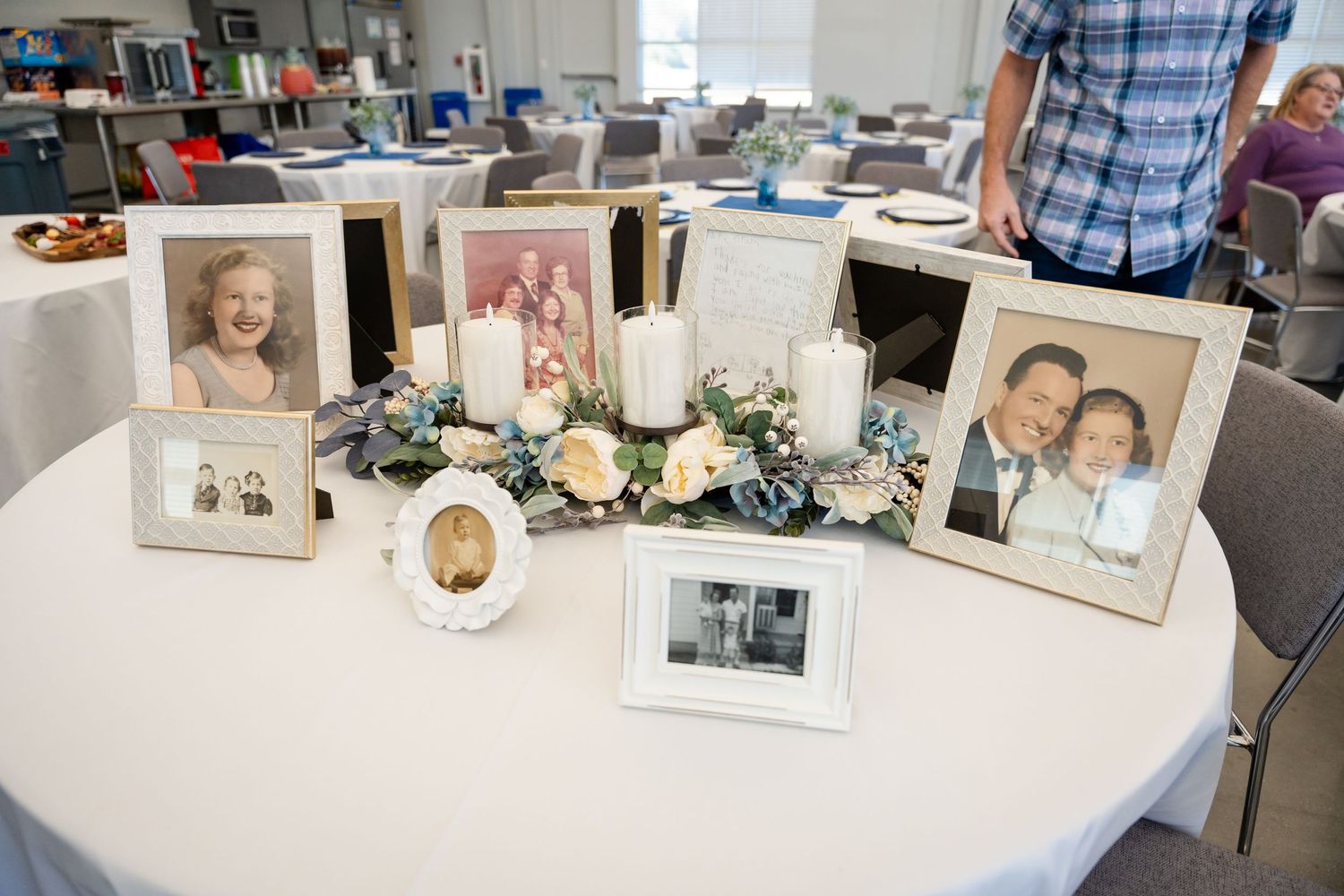 Memorial table display with vintage photographs, candles and floral arrangements on white tablecloth.