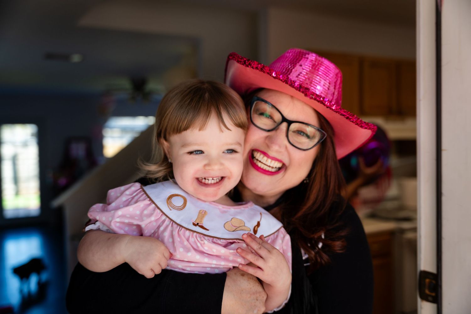 A pink-hatted woman in glasses shares a joyful moment with a small child wearing a pink dress.