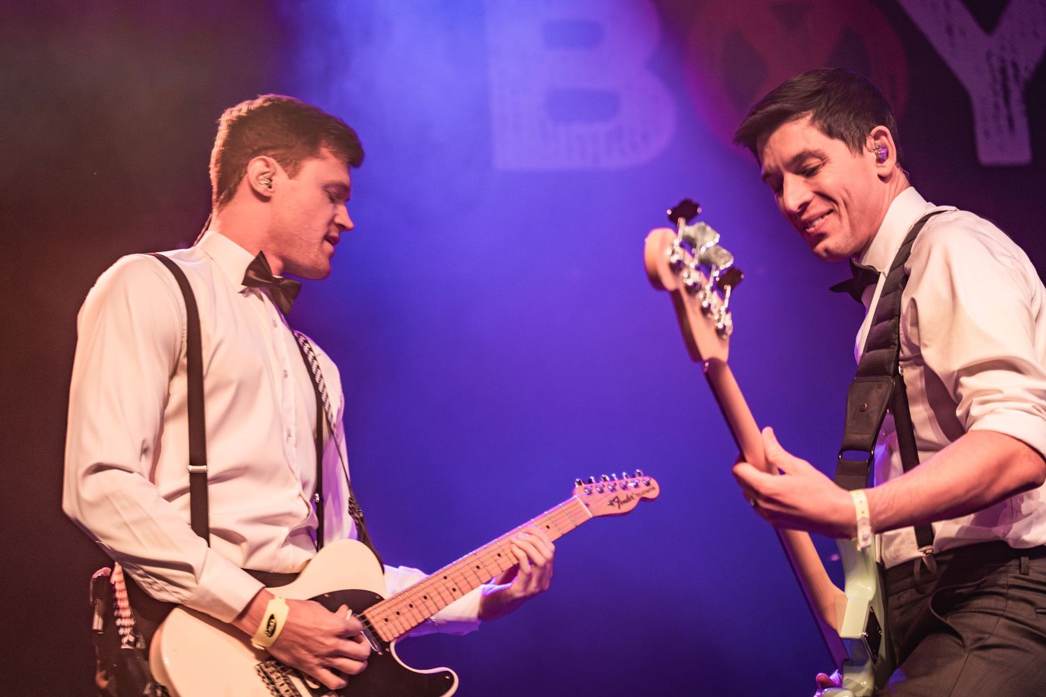 Two guitarists in matching white shirts and suspenders performing together on purple-lit stage.