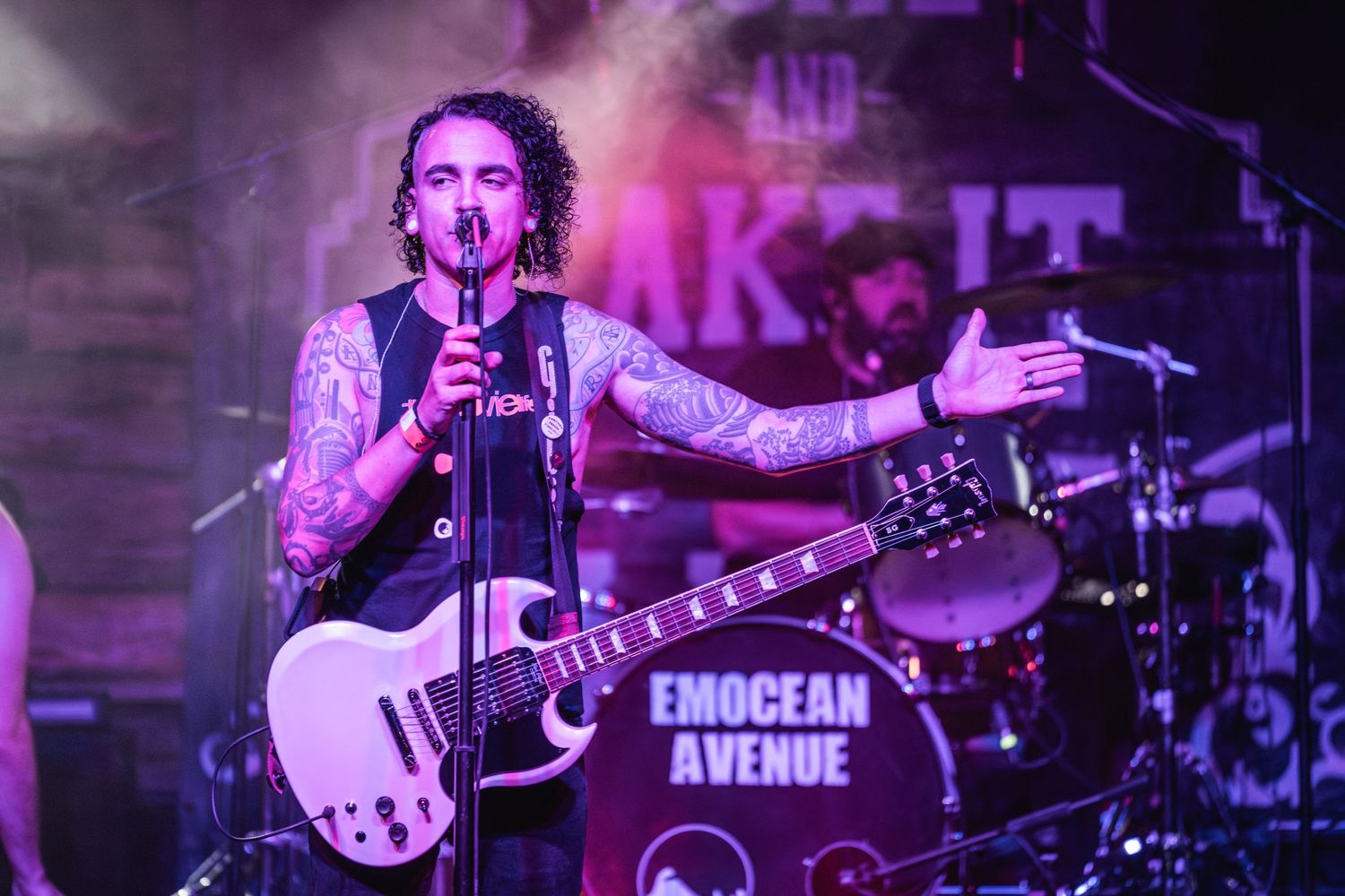 Musician with tattoos performs passionately with a white guitar under purple stage lights.