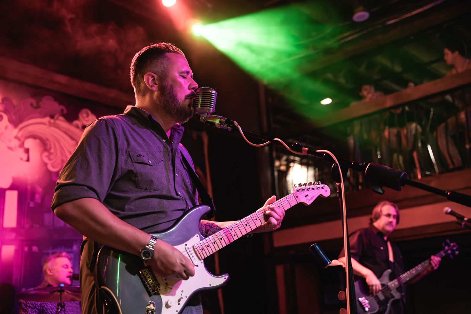 Musicians perform on stage with colorful lighting and vintage microphone setup.