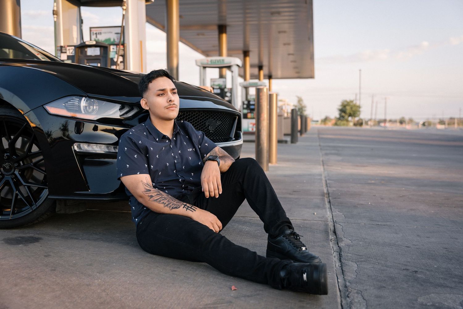Person in dark attire sitting beside black sports car at service station.