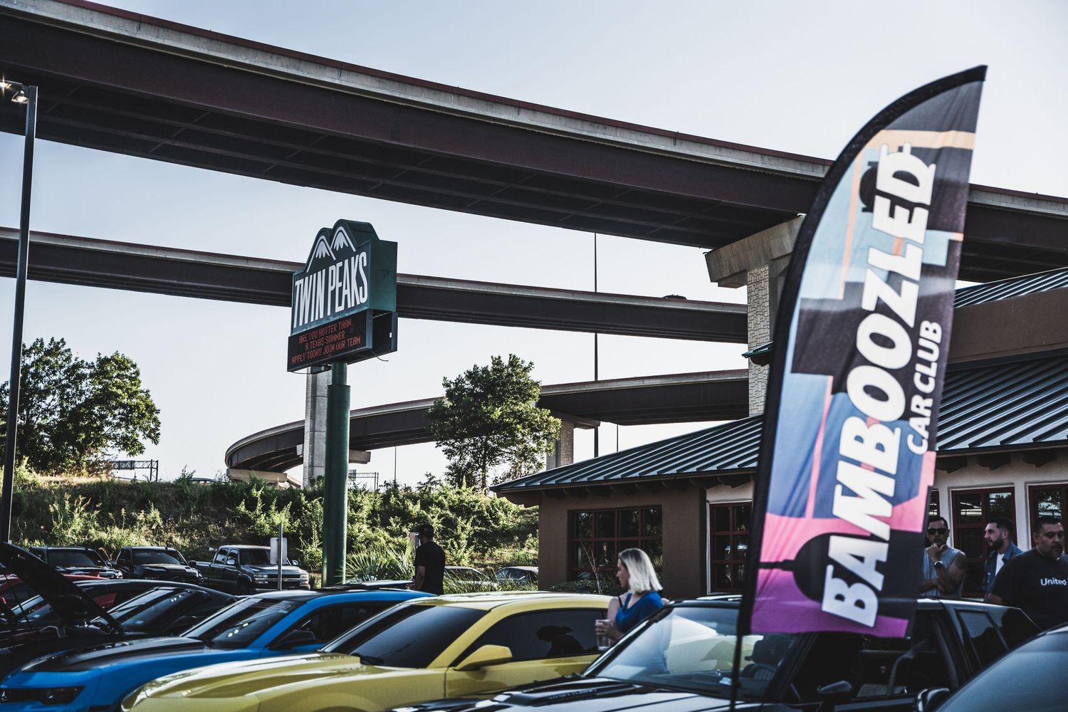 Car meet gathering at a retail location with colorful banners and multiple vehicles.