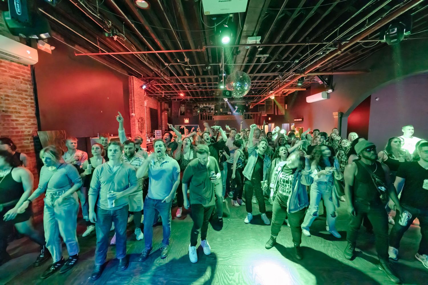 Large crowd dancing under green venue lighting with exposed industrial ceiling.