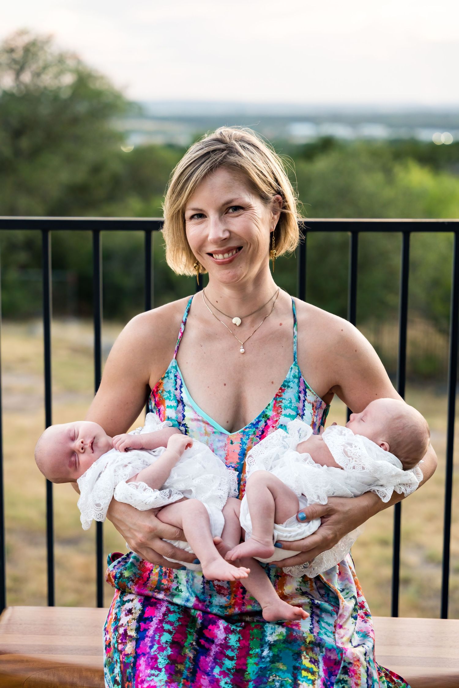 Woman in colorful dress holding twin babies on outdoor deck with scenic view.