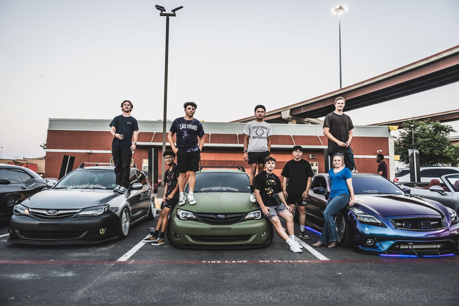Group of car enthusiasts gather around modified vehicles in parking lot under highway overpass.