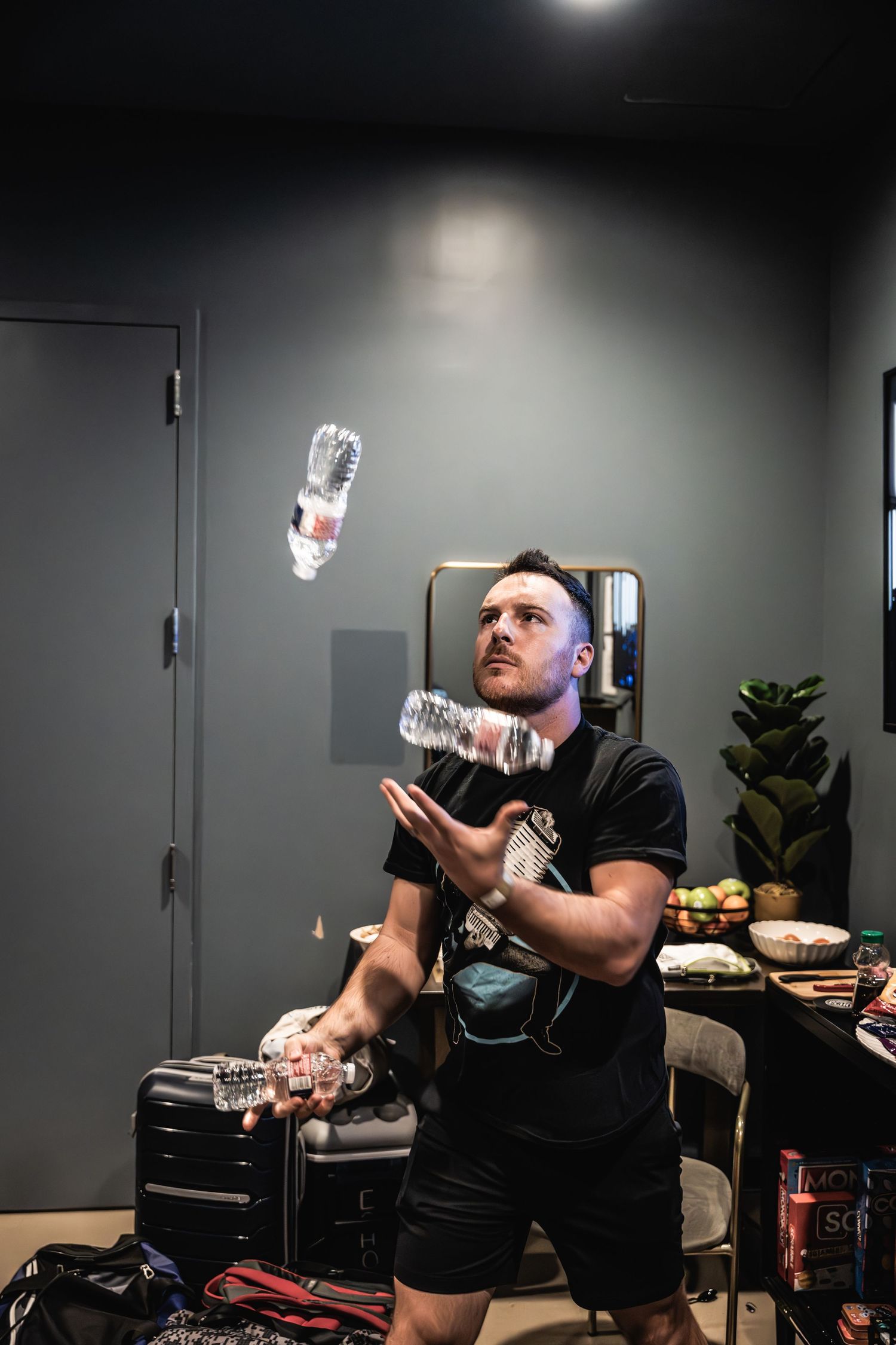 Person juggles water bottles backstage in a dark room with snacks and equipment visible.