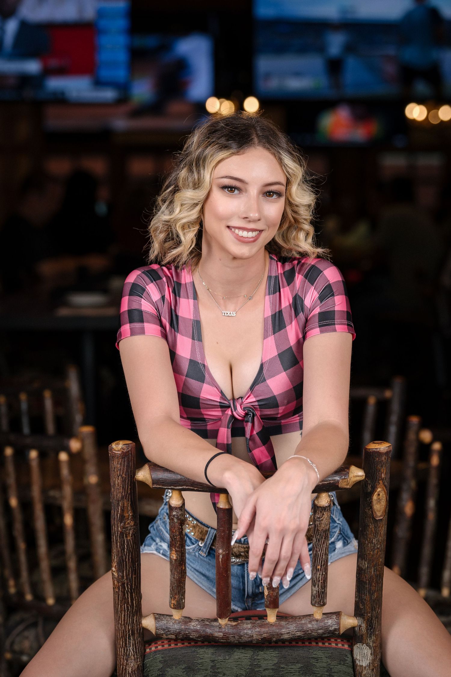 Model in pink buffalo check top and jean shorts sits on rustic wooden chair.