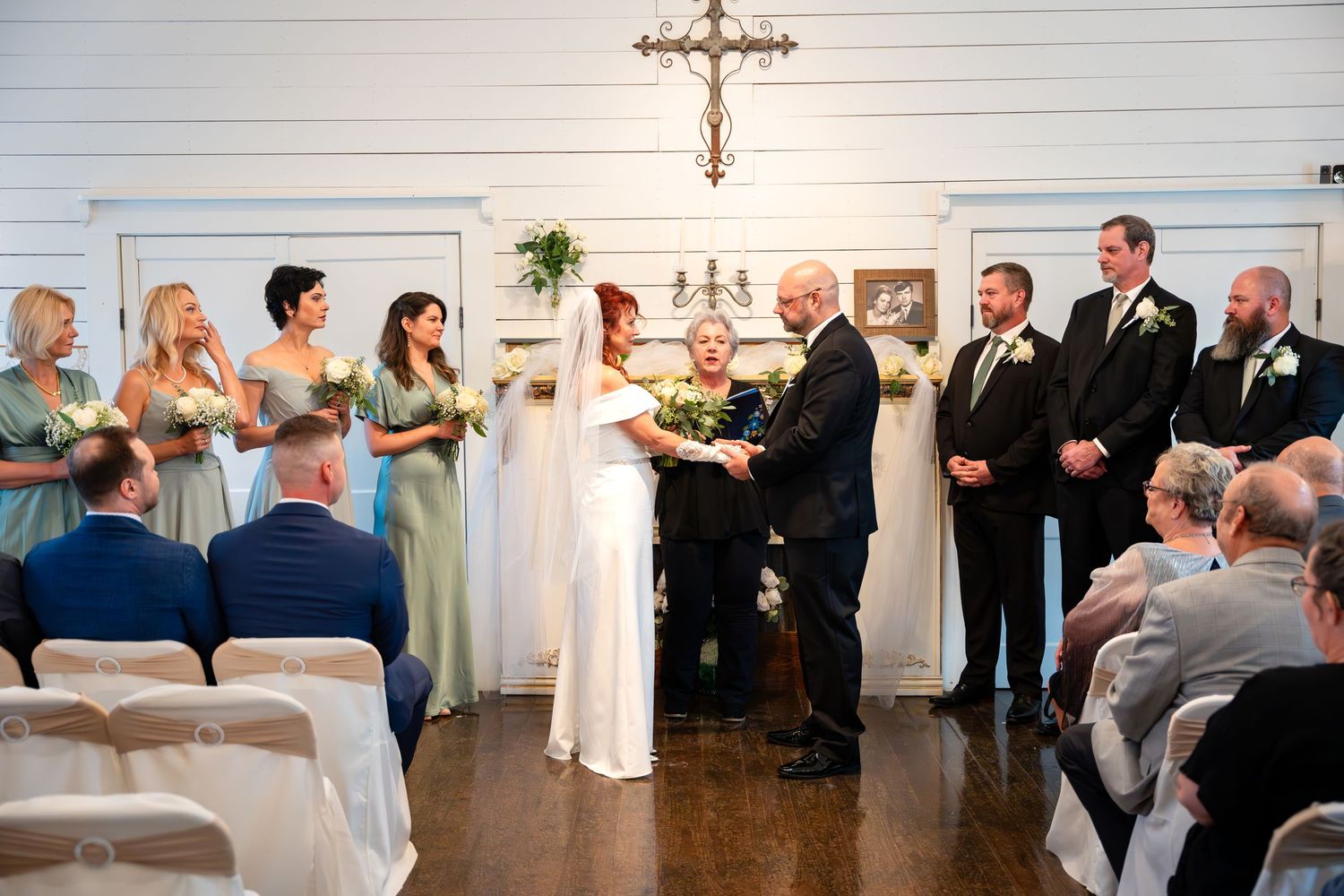 Wedding ceremony in white chapel with bridal party in sage green dresses and dark suits.