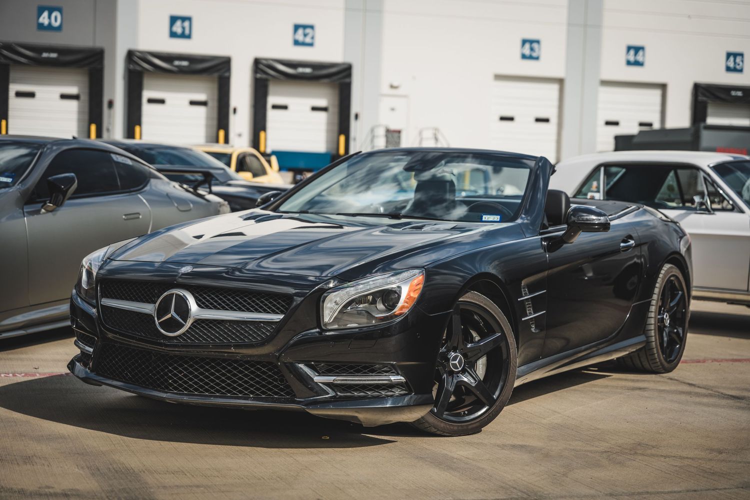 Black Mercedes-Benz SL convertible with dark wheels parked in front of industrial loading bays.