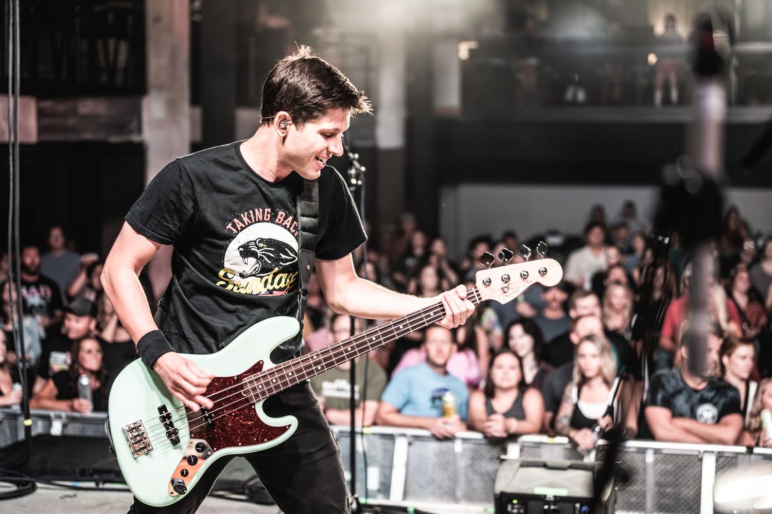 Bass guitarist performs energetically on stage during a concert with crowd visible in background.