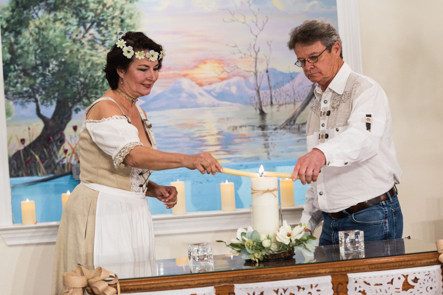 Couple performing candle lighting ceremony against painted backdrop of sunset over water.