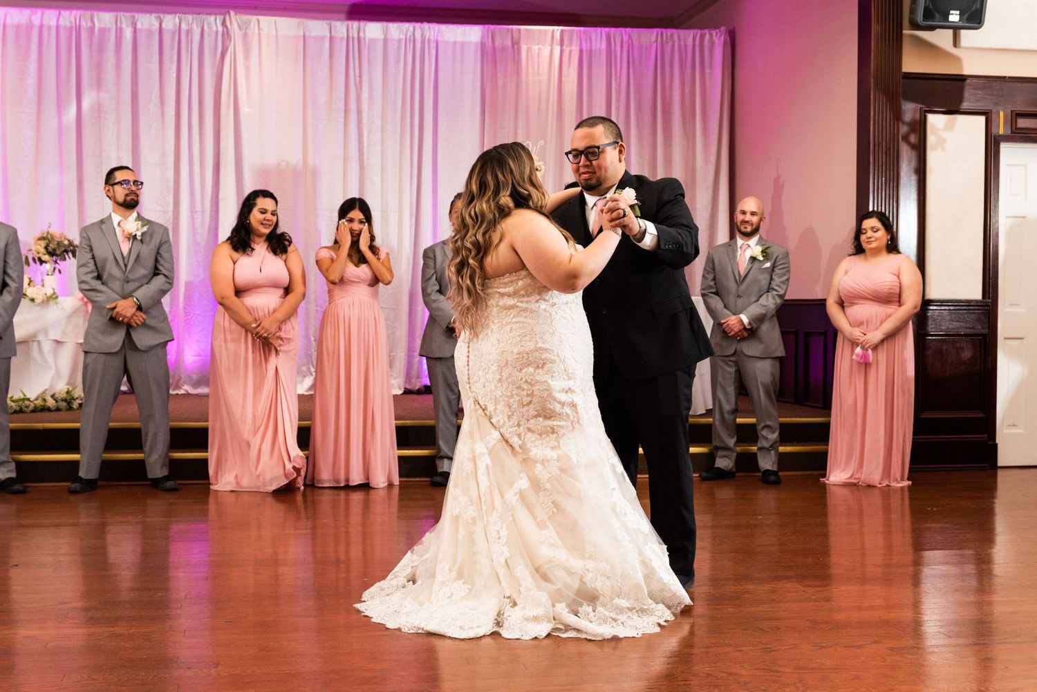Wedding party watching couple's first dance in reception hall with pink backdrop.