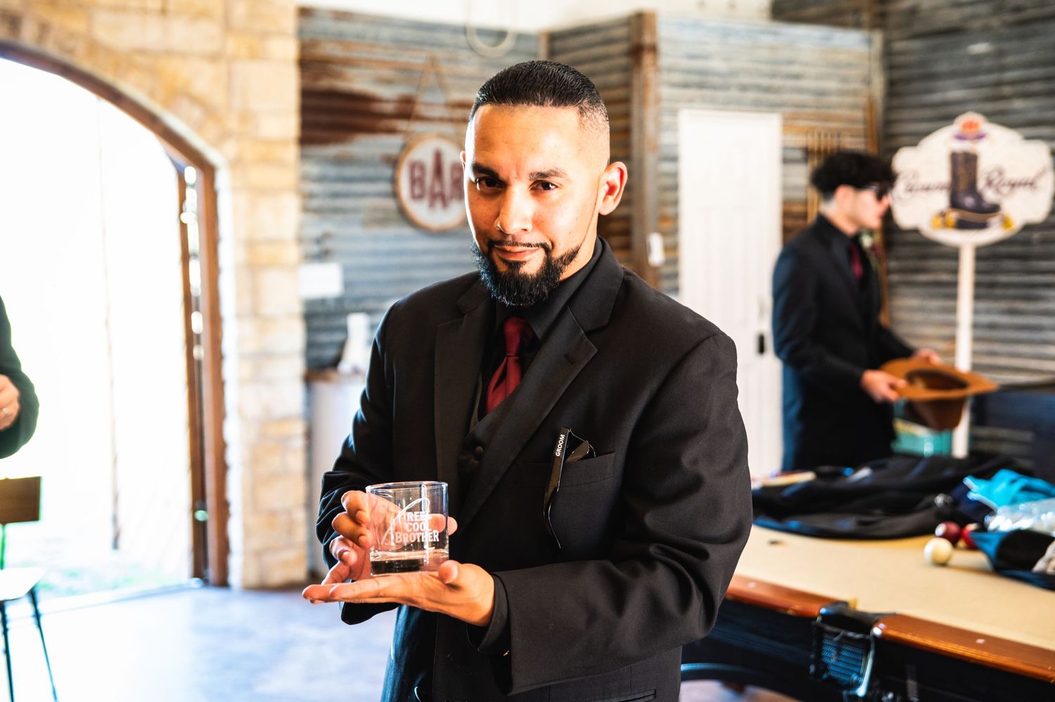 A person in a black suit holds a small glass in an indoor setting.