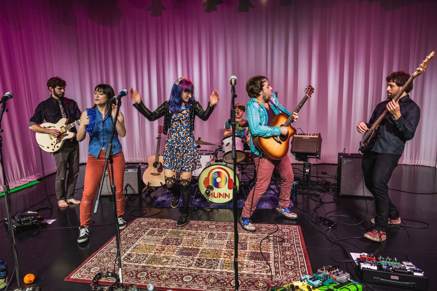 Band members play various instruments on stage with oriental rug and pink curtains in background.