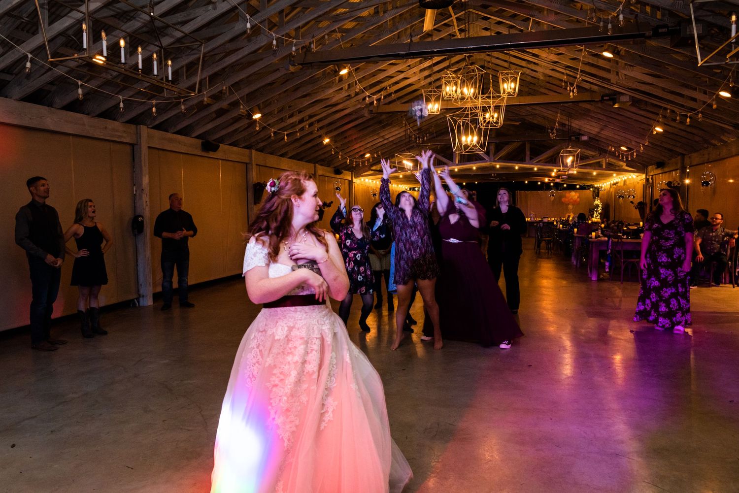 Bride in a pink-lit reception venue stands under rustic wooden beams and string lights.