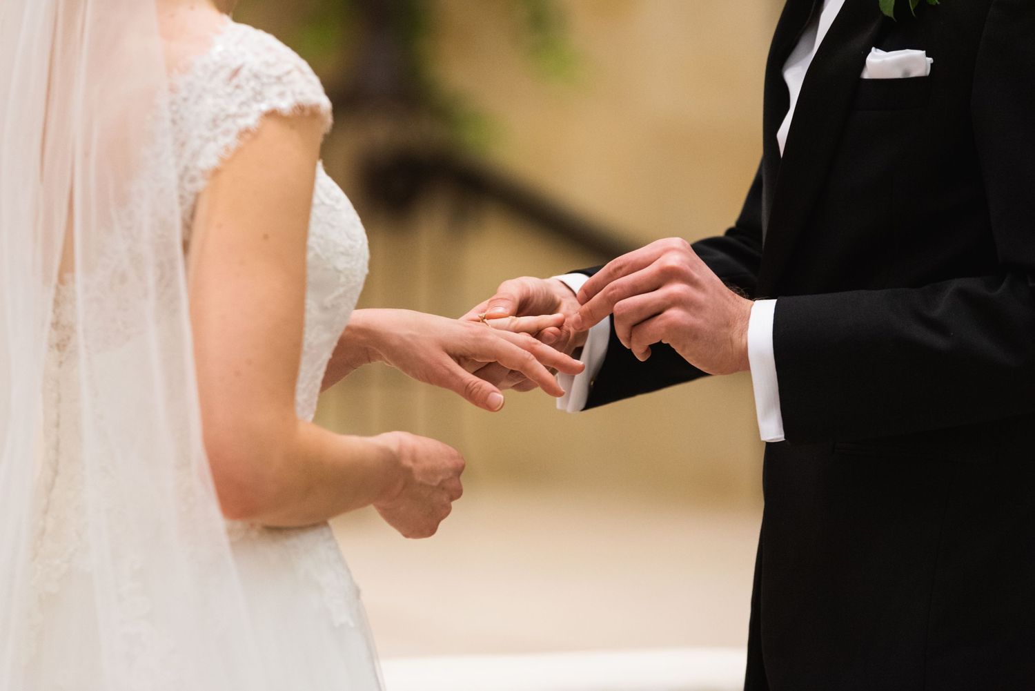 Close-up of wedding ring exchange during ceremony with bride in white lace gown and groom in black suit.