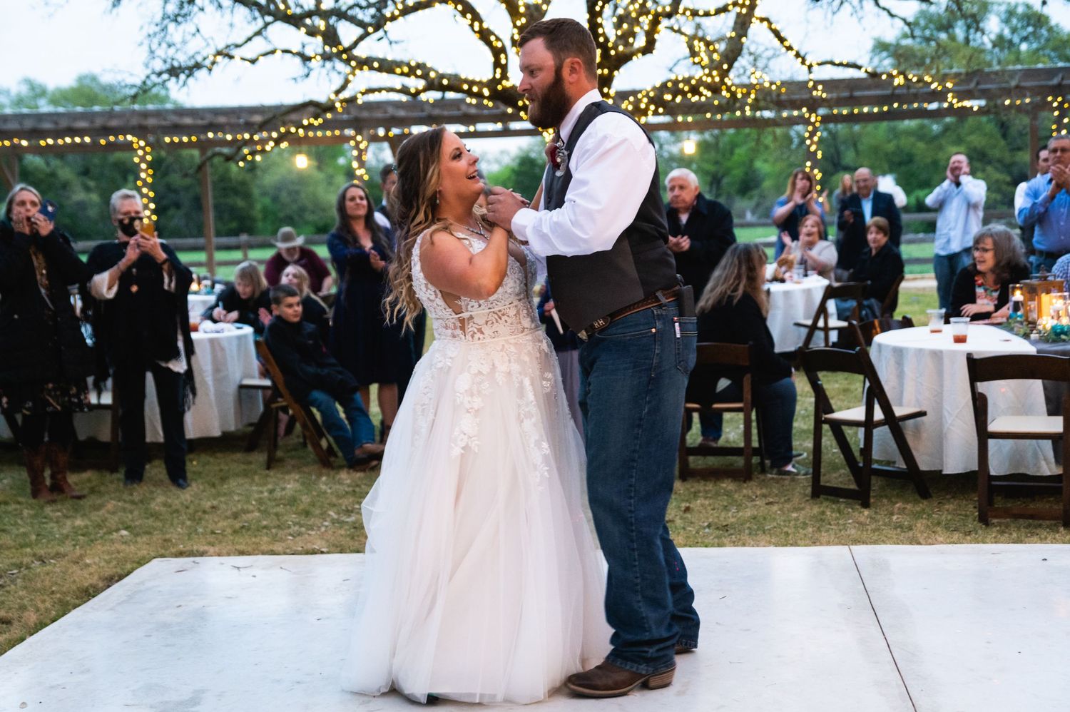 A couple dances on a white platform under twinkling lights at an outdoor wedding reception.