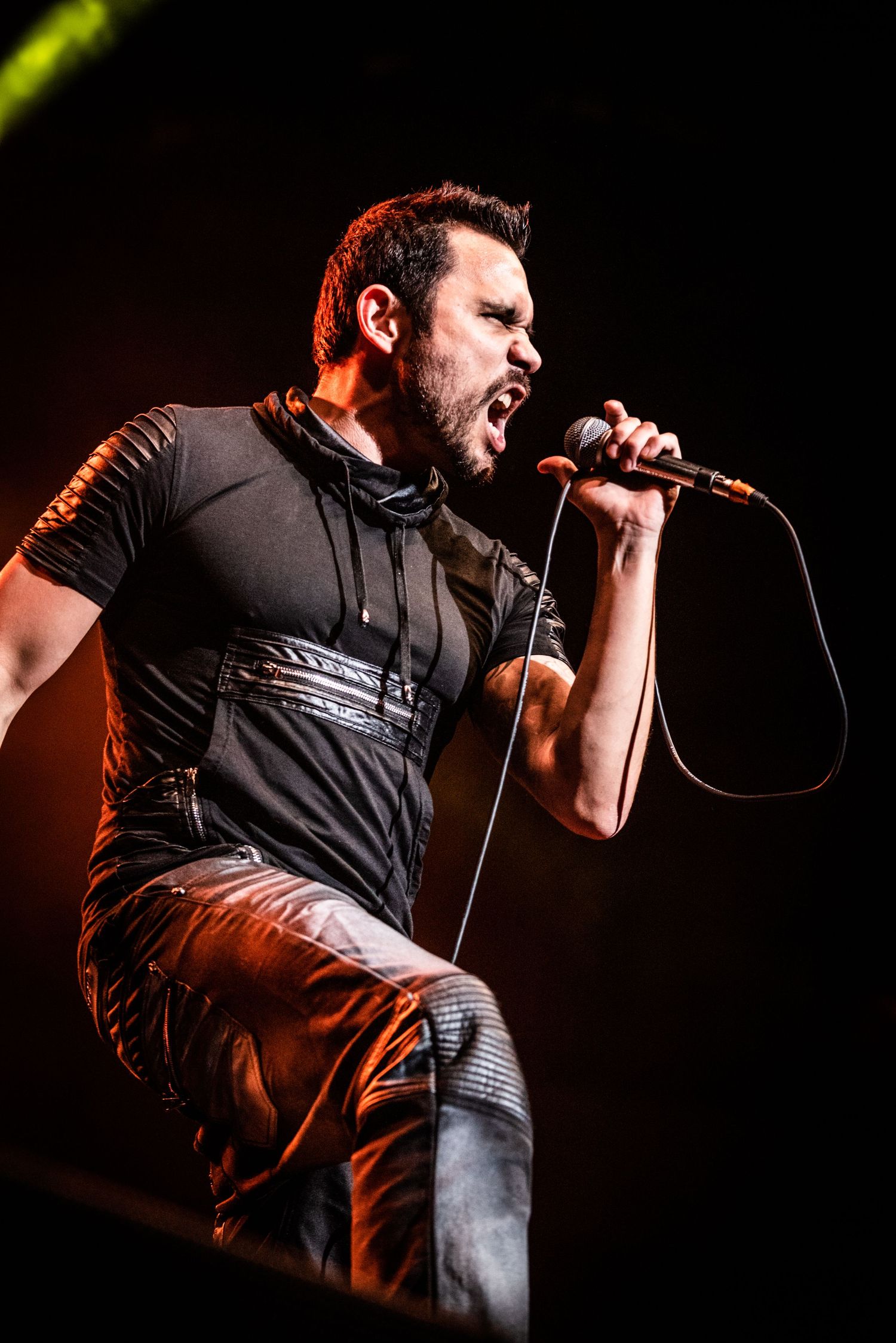 A rock vocalist performs passionately on stage in a black outfit under dramatic lighting.