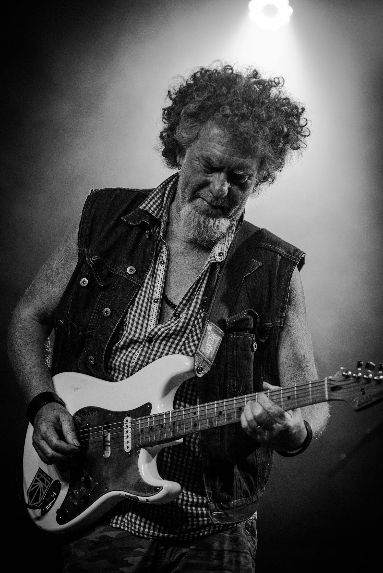 Concert performer plays electric guitar while wearing checkered shirt and denim vest under stage lights.