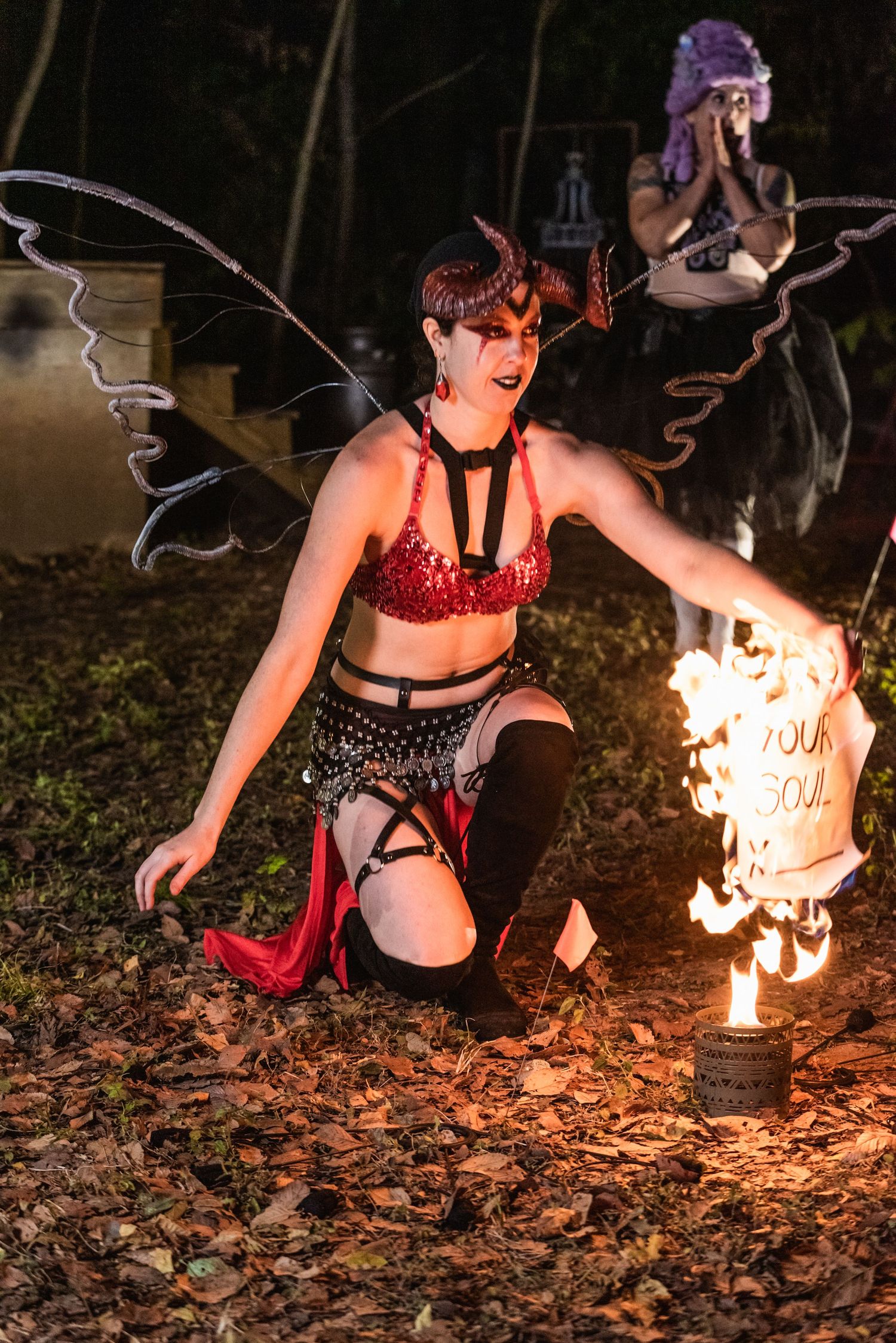 Performer in dark red and black costume kneels dramatically near flaming prop during outdoor night performance.