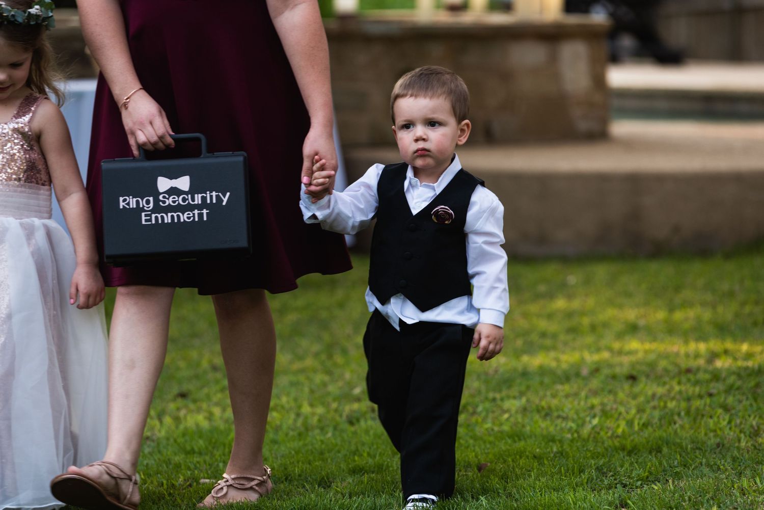 A small child in formal attire walks on grass beside someone holding a ring bearer sign.