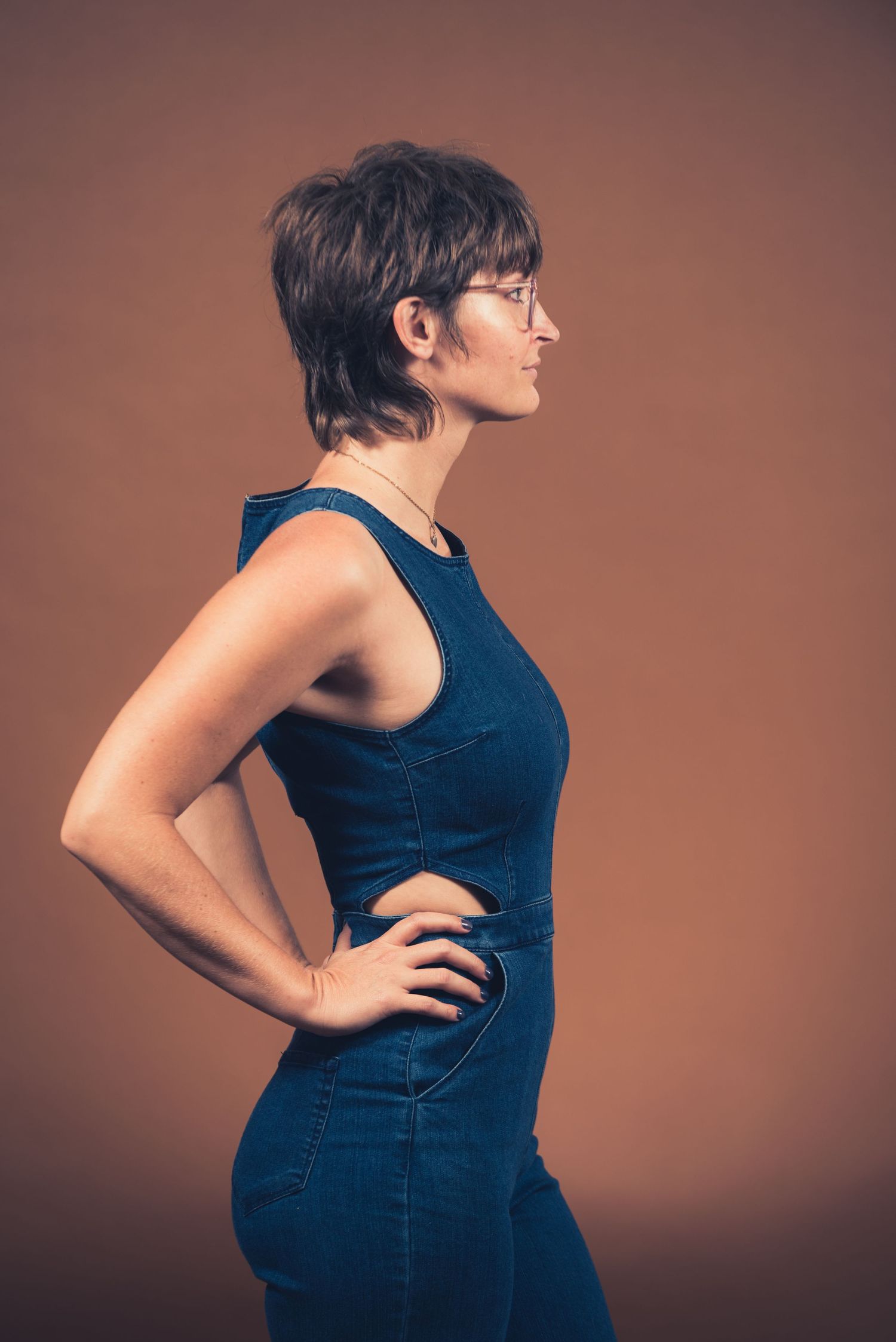 Side profile portrait of someone in a navy blue sleeveless outfit against a brown backdrop.