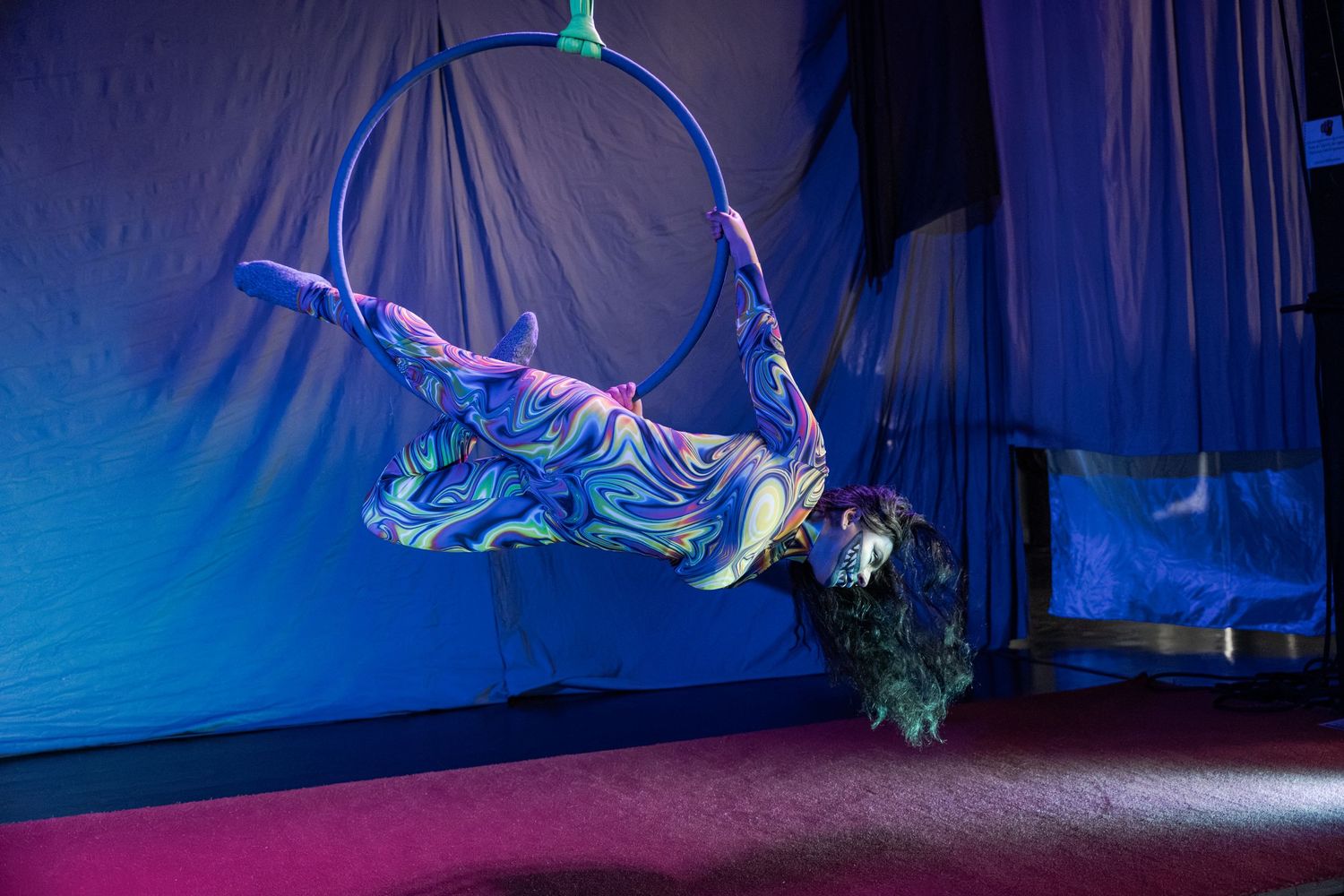 Aerial hoop performer in colorful costume gracefully balancing inside a lyra ring under blue stage lighting.