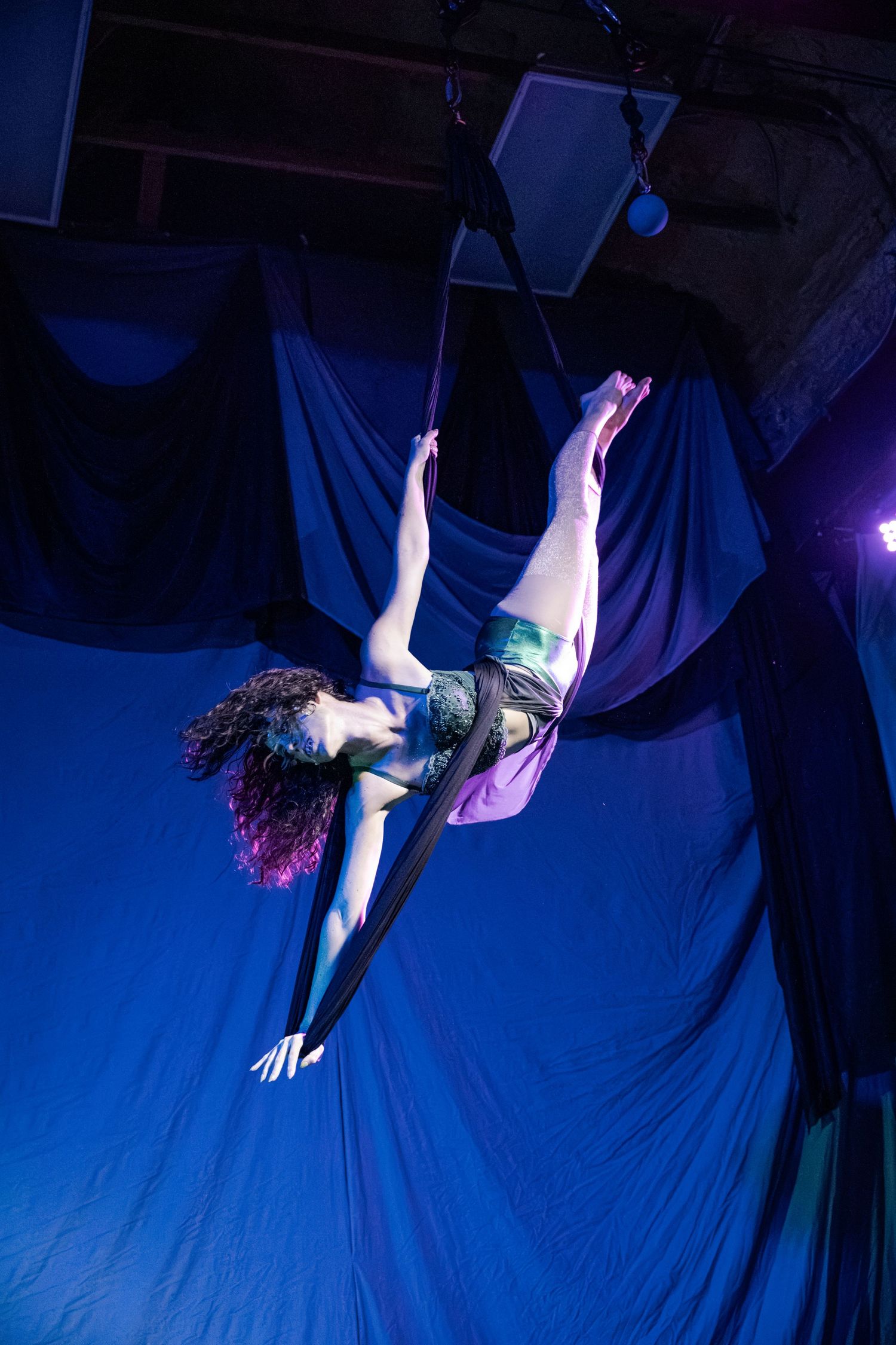 Aerial acrobat performs graceful inverted pose on silk fabric under blue stage lighting.