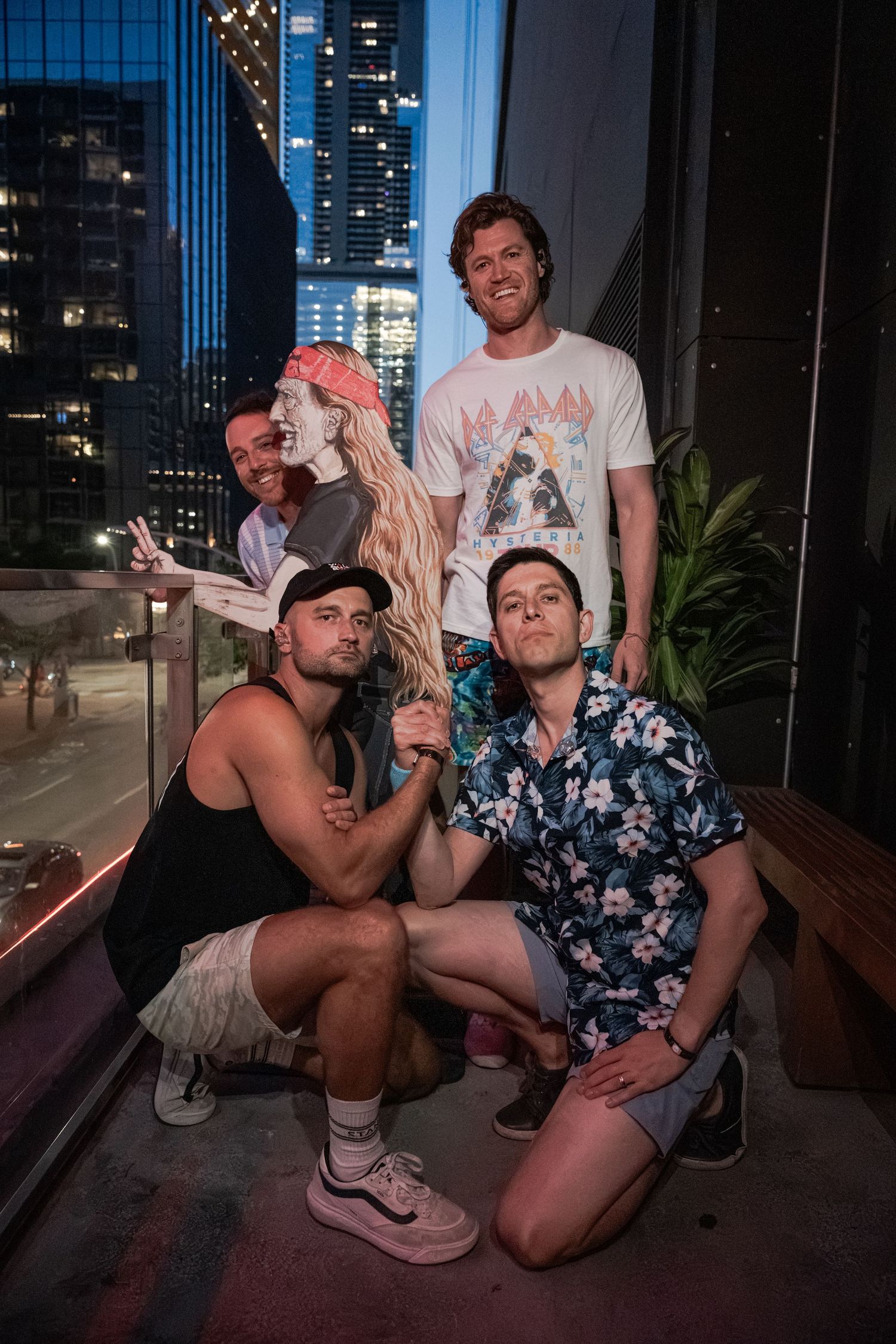 Group of five friends posing on a rooftop balcony at night with city skyline in the background.