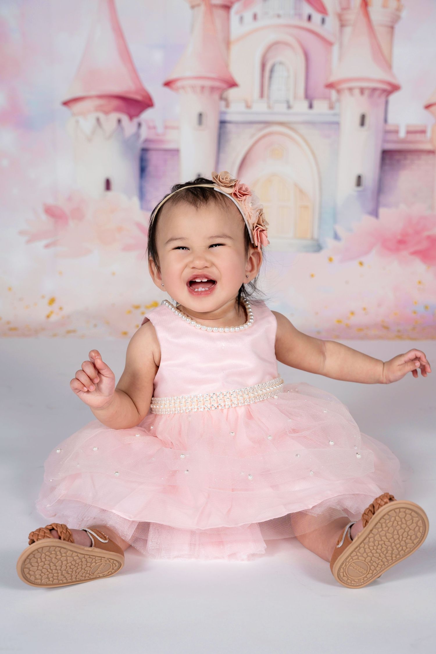A laughing baby girl in a pink tutu dress sits before a fairy tale castle backdrop.