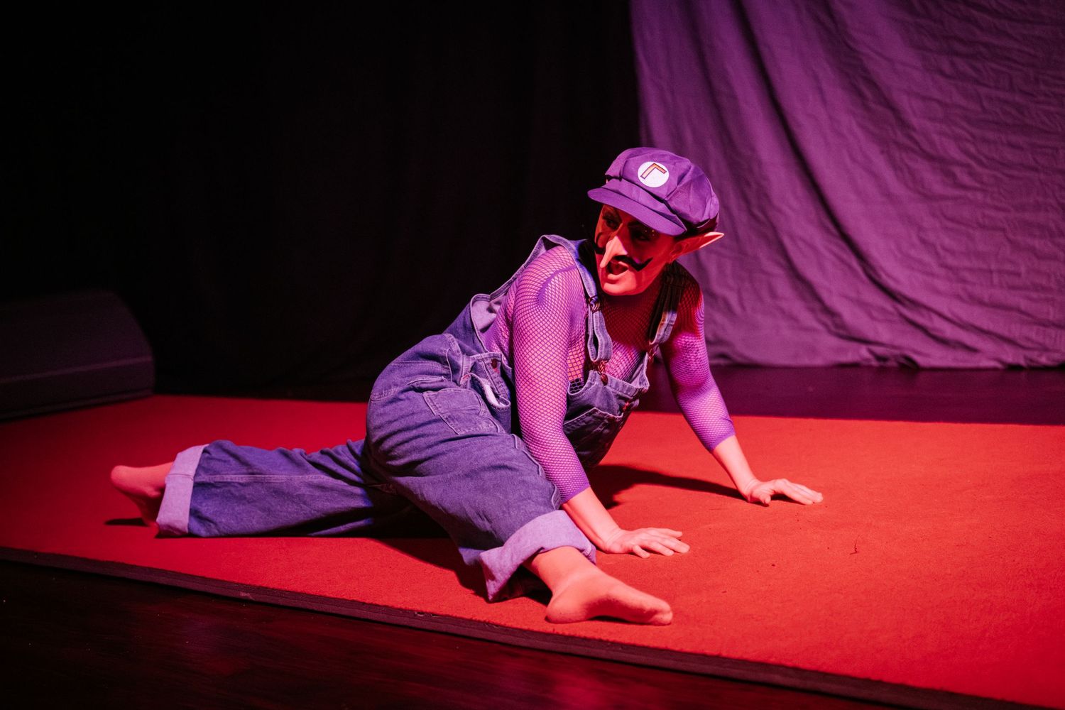 Performer in purple overalls and cap crawls on red stage floor under dramatic lighting during theatrical performance.
