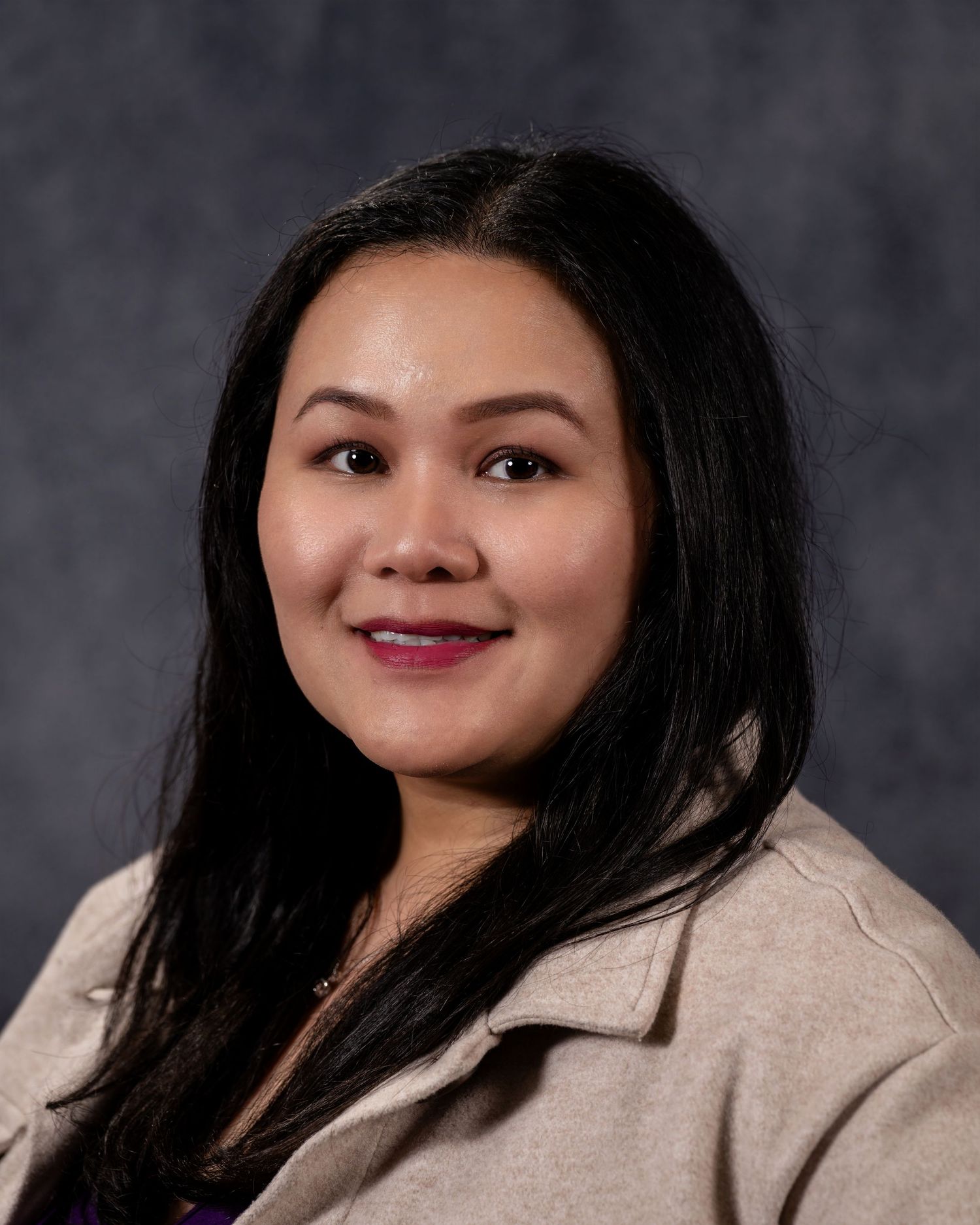 Professional headshot of a smiling woman with long black hair wearing a beige blazer against a gray background.