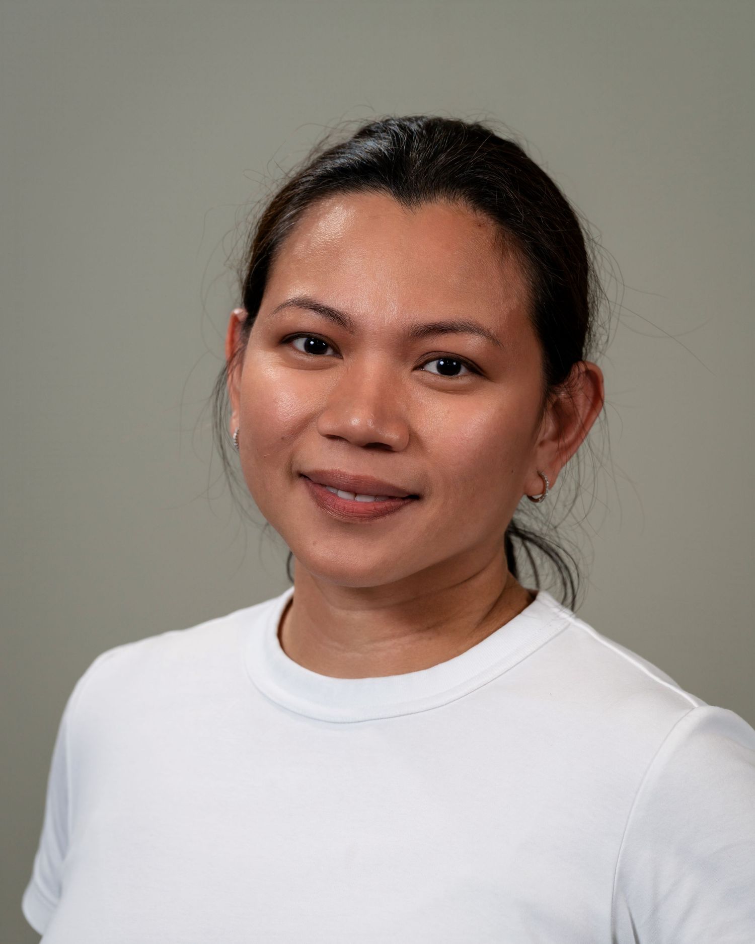 Smiling woman with dark hair wearing a white crew-neck shirt posed against a neutral gray background.