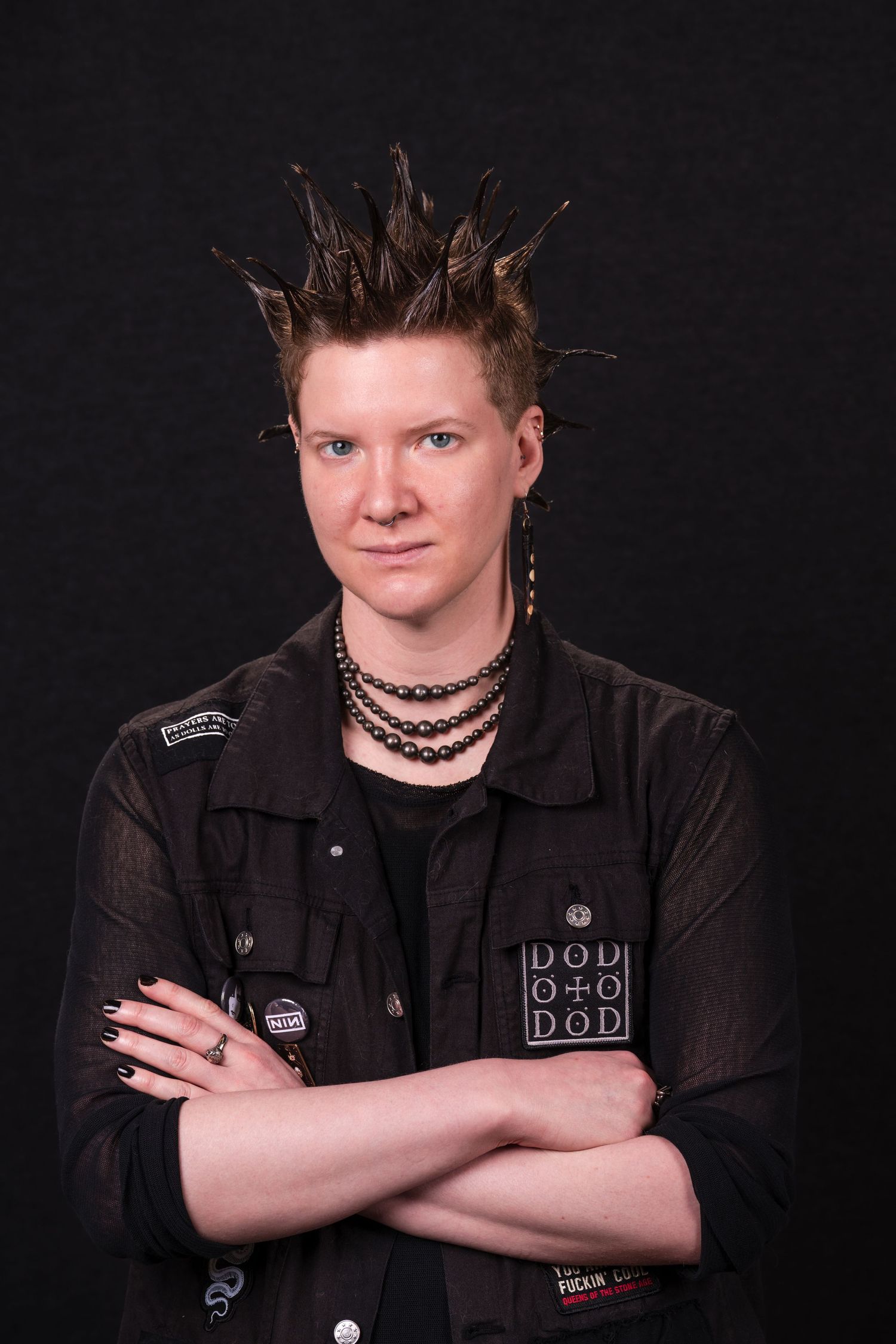 Portrait of someone with spiked hair wearing a black jacket and necklaces against a dark background.