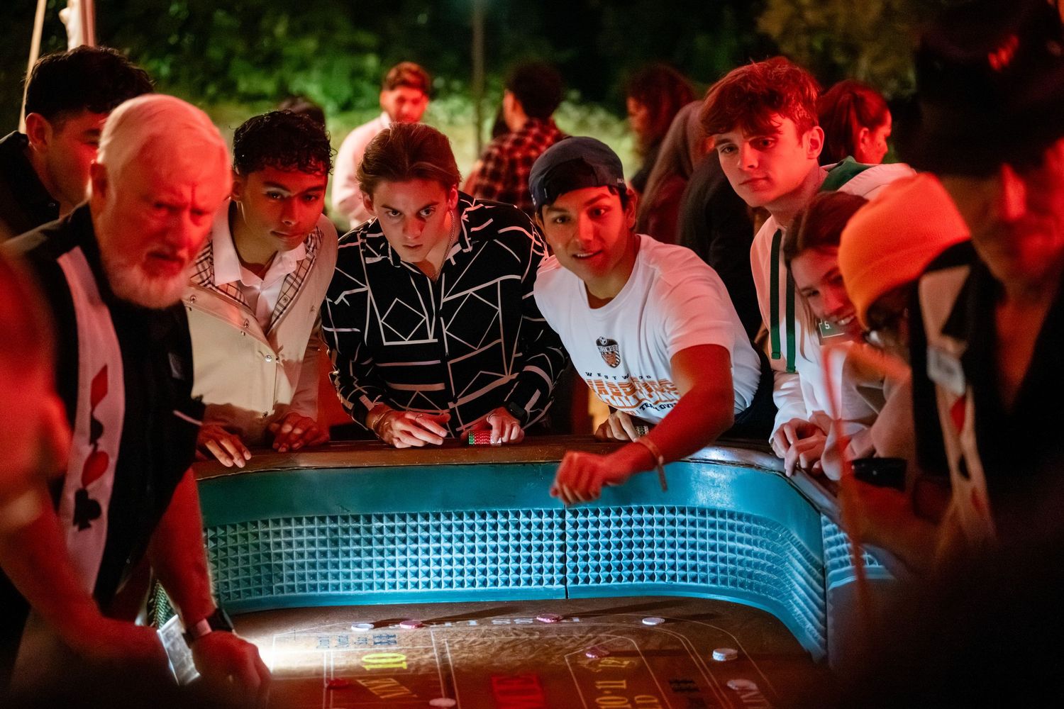 People gather around illuminated poker tables in a dimly lit casino environment with red ambient lighting.